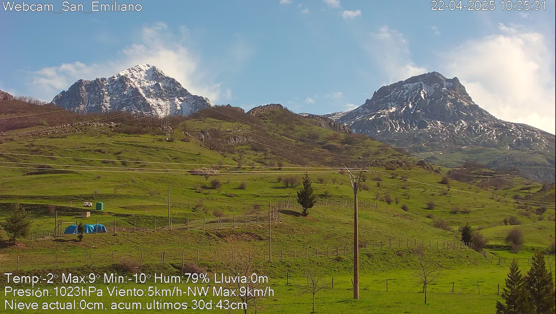 Two snow-capped mountains rise in the distance over a grassy valley with utility poles and fences under a mostly clear, sunny sky.