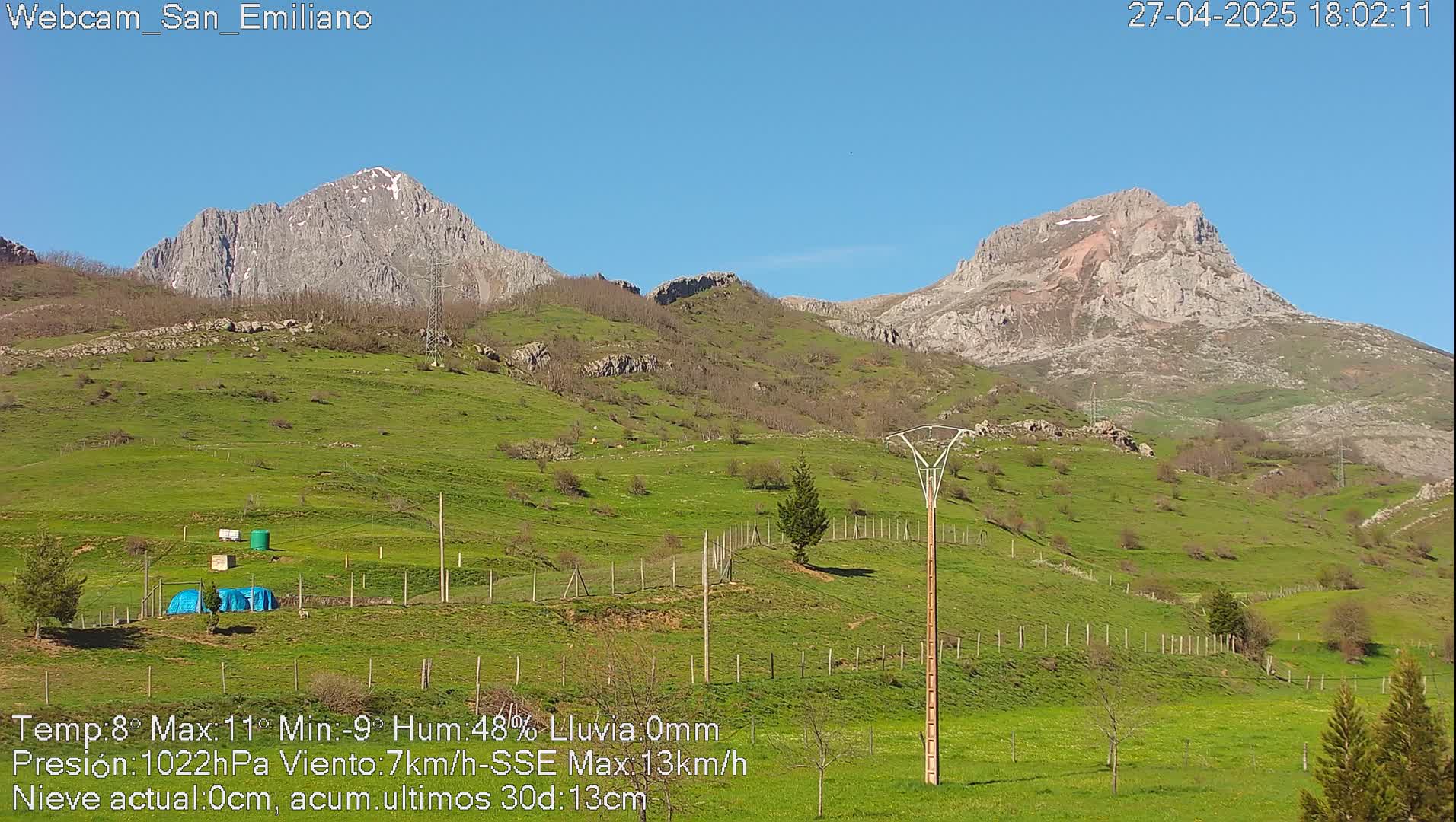 A sunny day reveals rolling green hills and mountains with some snow-capped peaks under a clear blue sky.