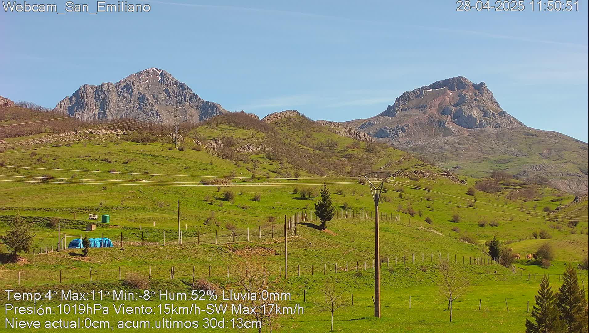 Two mountainous peaks rise in the background of a sunny, green valley dotted with small trees and fences under a clear blue sky.