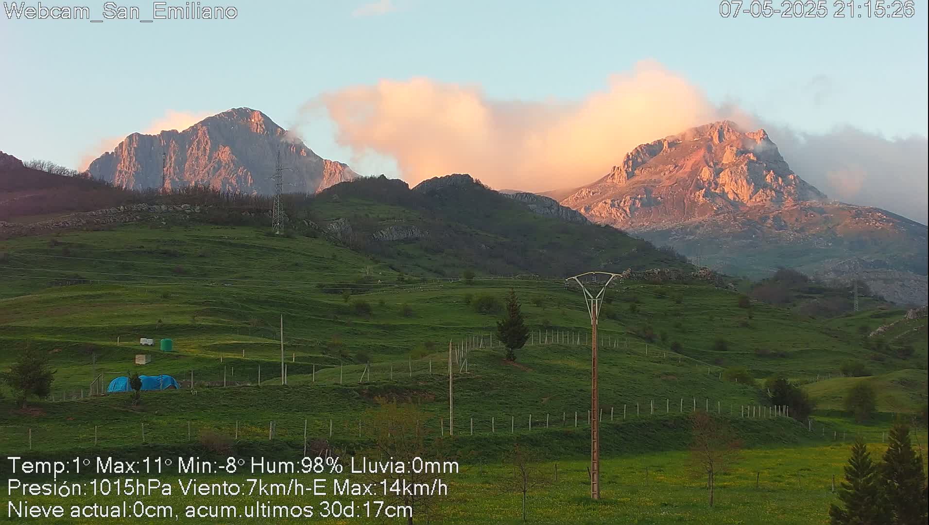 Rolling green hills at sunset under a partly cloudy sky, with two large mountains in the background.