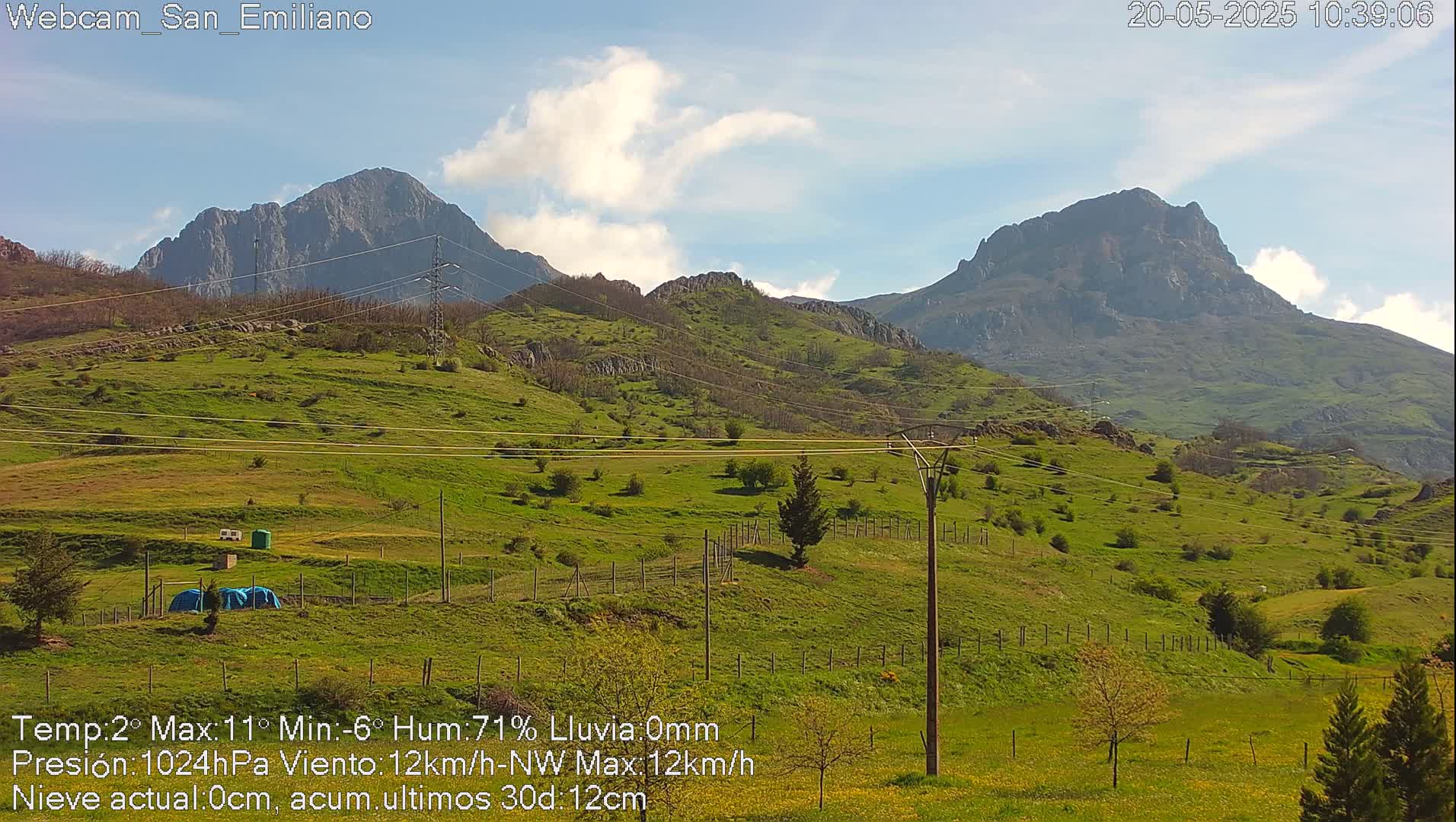 A partly cloudy day reveals two mountainous peaks in the distance, overlooking a green hillside dotted with small trees and power lines.