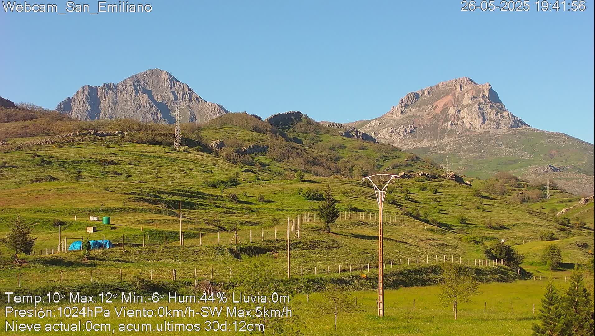 Two rocky mountains rise in the background of a grassy hillside under a clear blue sky.