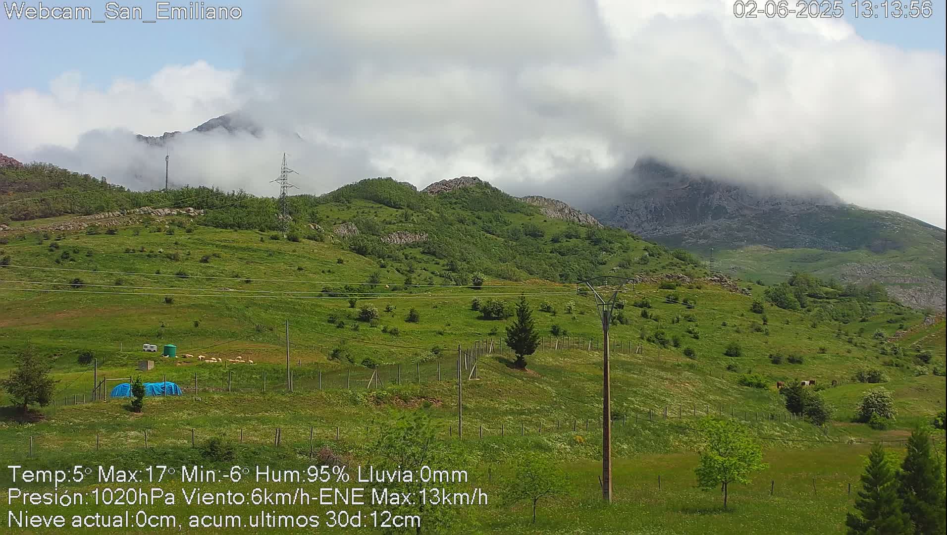 A partly cloudy day reveals green hills and mountains partially obscured by clouds, with livestock grazing in a fenced pasture.