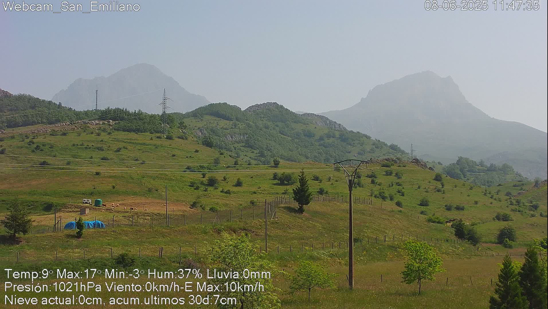 A hazy, sunny view of rolling green hills and distant mountains, with some power lines and sparse vegetation in the foreground.