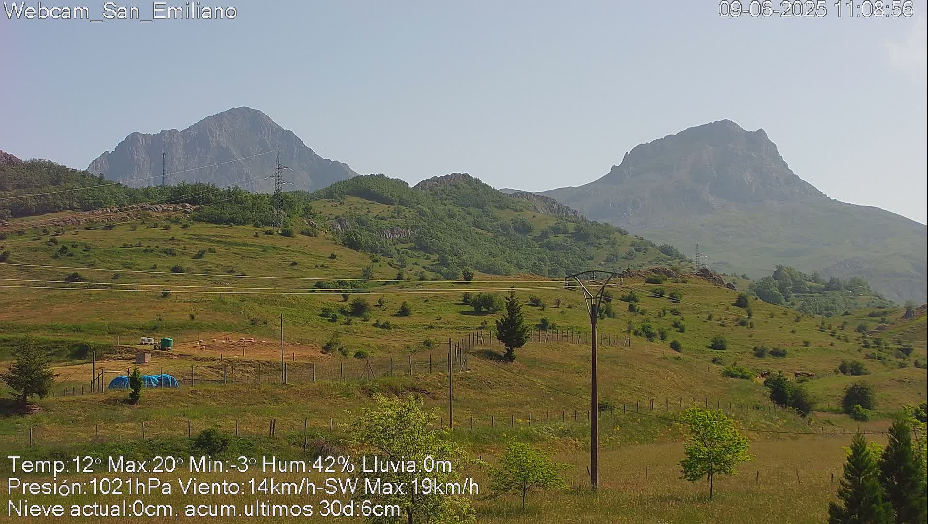 A grassy hillside with a fence, power lines, and small structures is visible in front of two large, distant mountains under a clear, sunny sky.