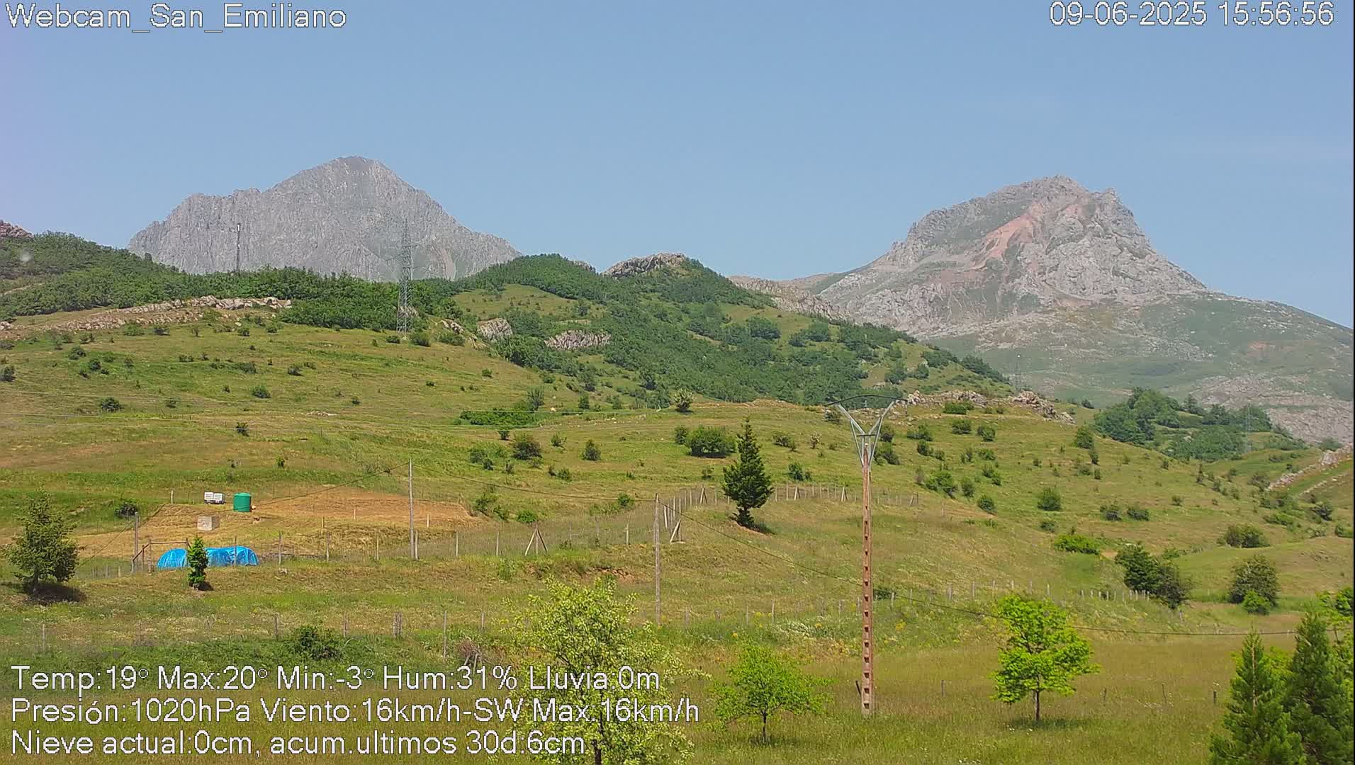 Two rocky mountains rise in the background of a sunny, grassy landscape dotted with small trees and utility poles.