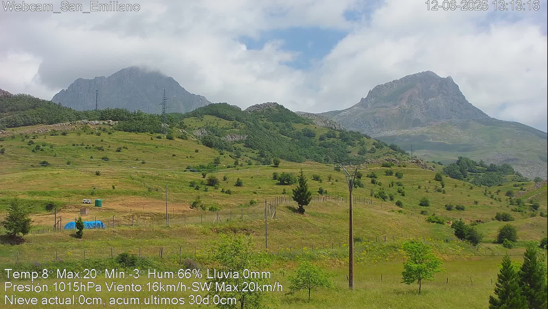 A partly cloudy day reveals rolling green hills dotted with trees and power lines, with large mountains in the background.