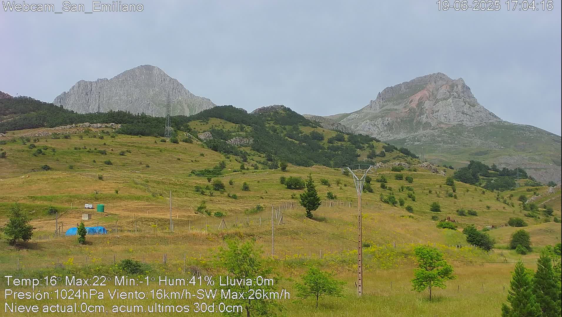 A mountainous landscape under an overcast sky shows rolling green hills dotted with small trees and shrubs, with two prominent, rocky peaks in the background.