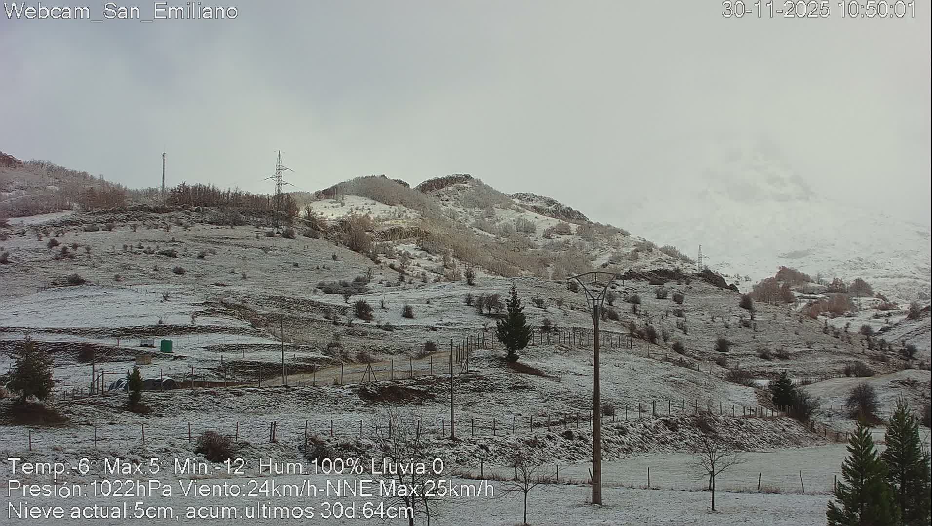 A snow-dusted mountain landscape with bare trees, fences, and power lines stretches under a grey, overcast sky.
