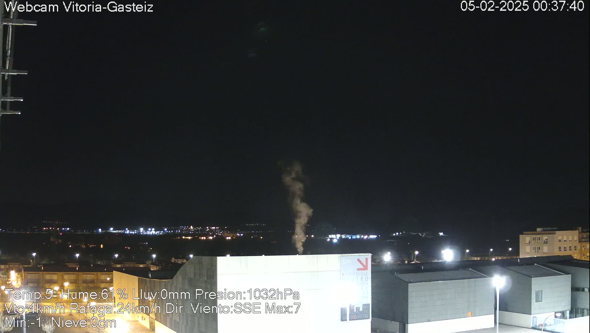 A plume of smoke rises into a dark night sky over a city illuminated by lights.