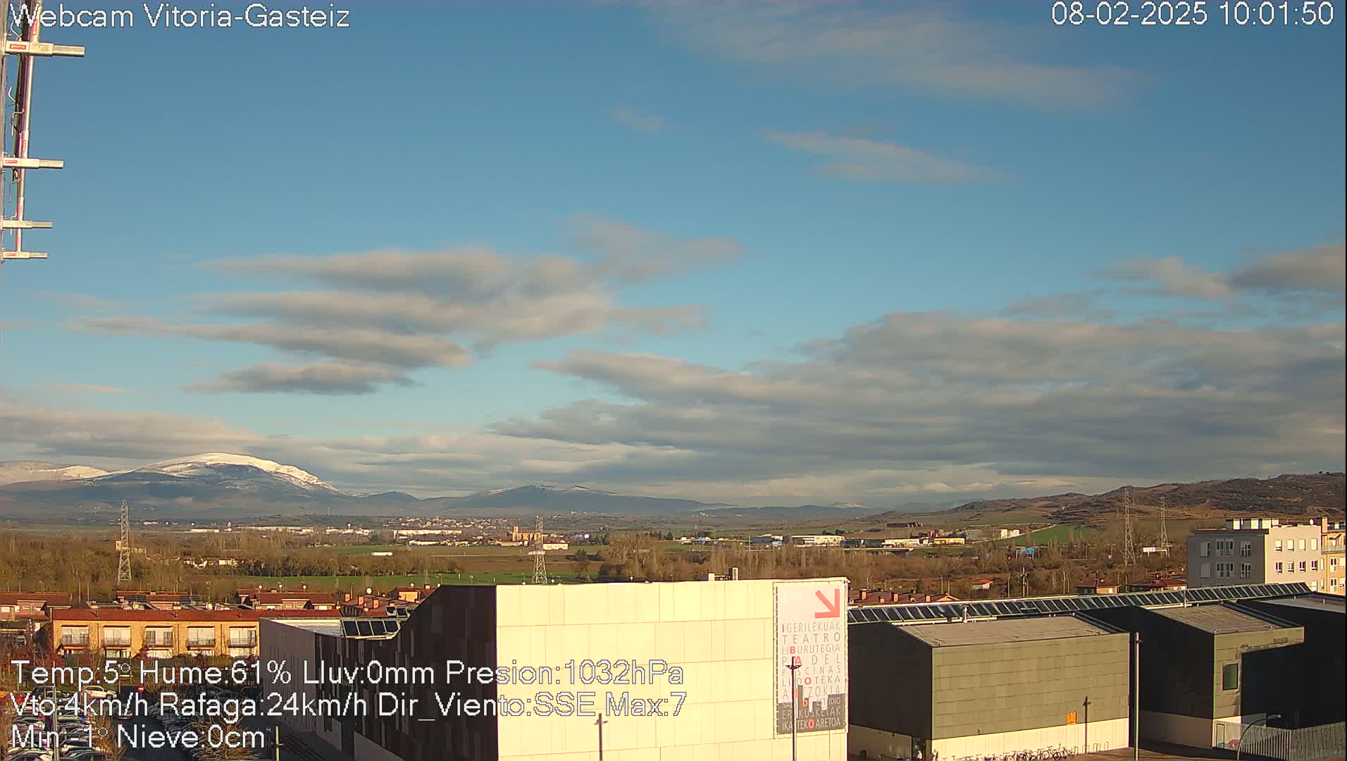 A partly cloudy daytime scene shows a cityscape with buildings and mountains in the distance, the mountains having some snow on their peaks.