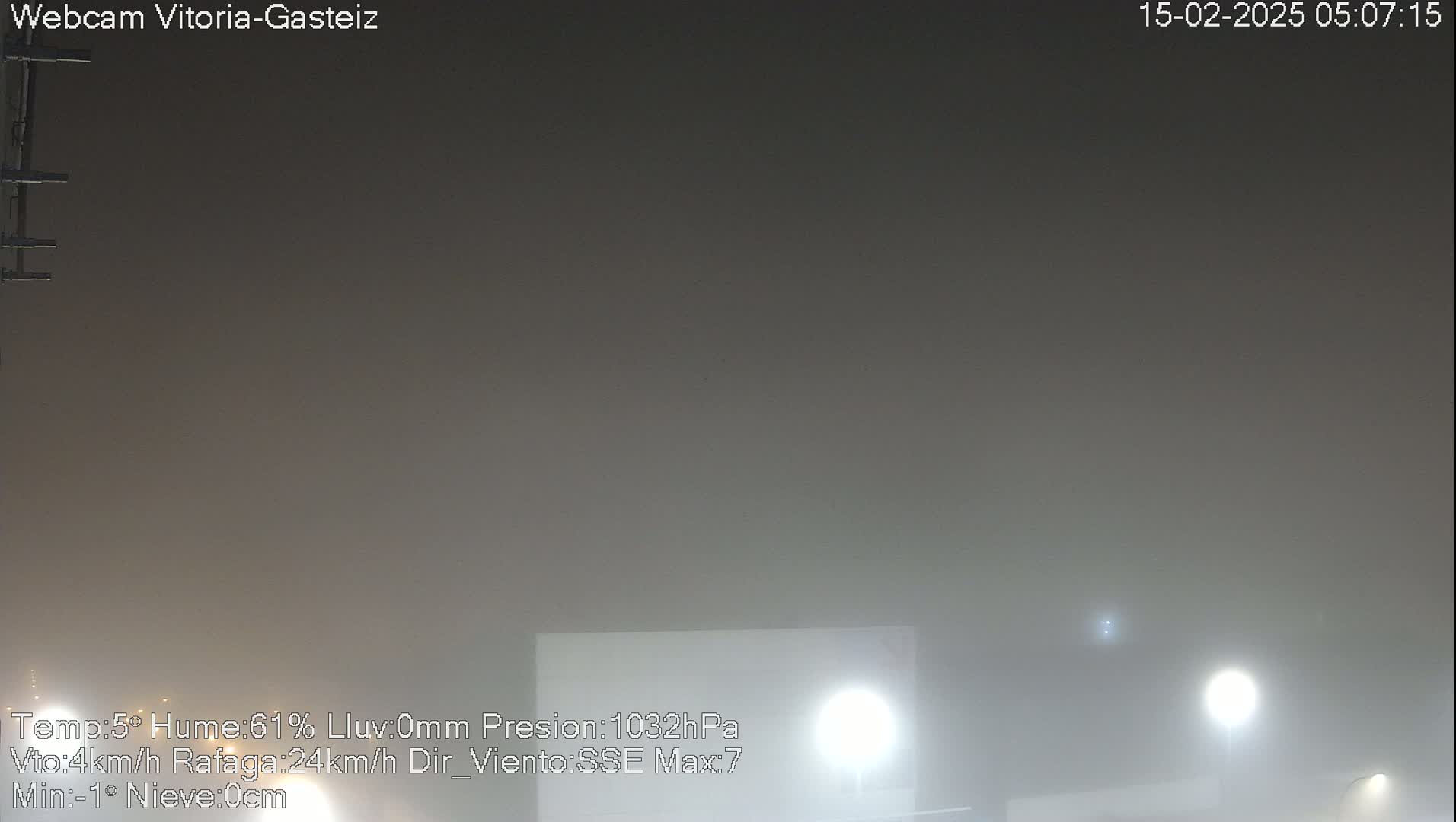 A thick fog obscures the view of a dimly lit area at night, with only a few lights visible.