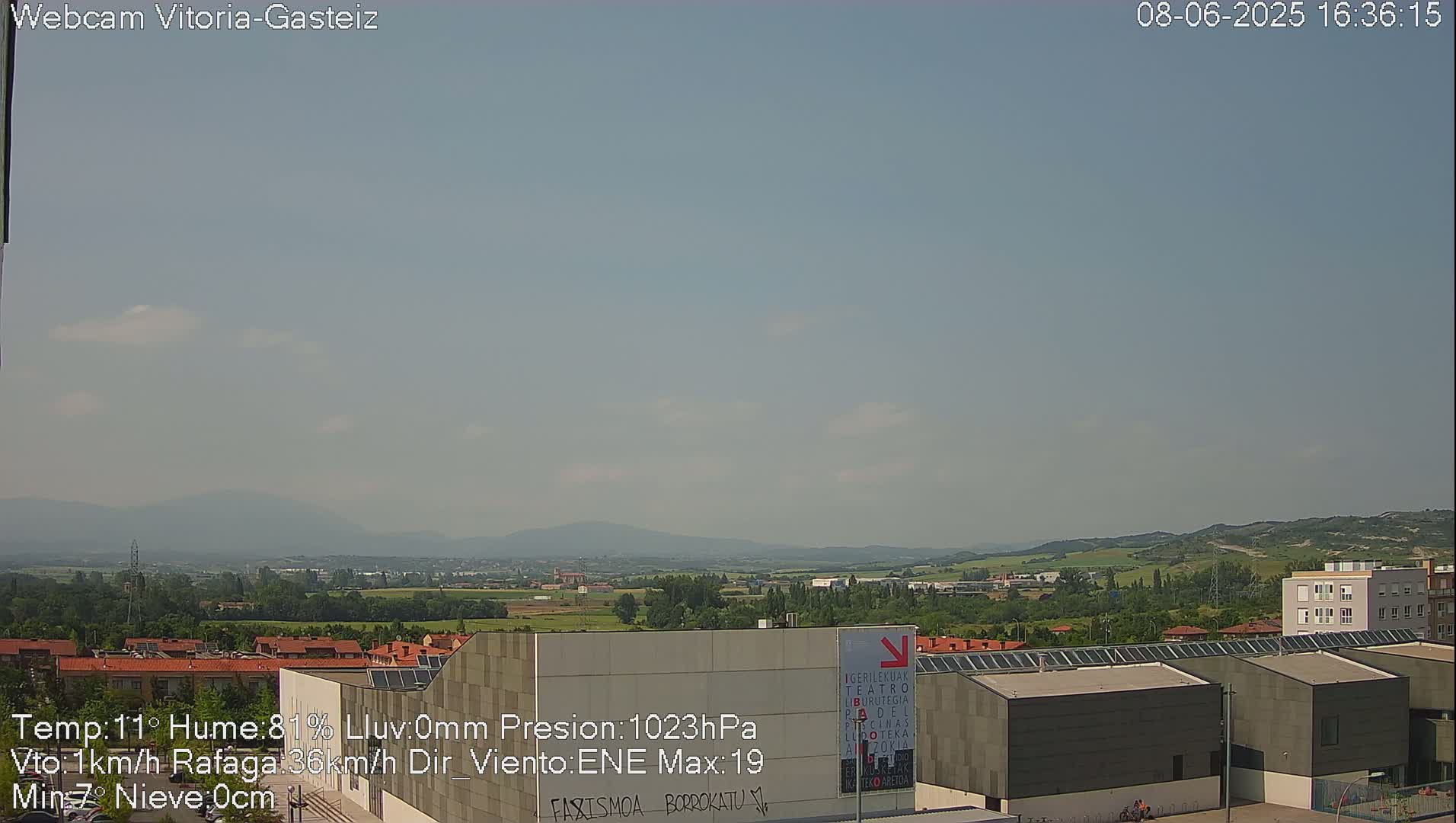 A hazy, sunny day reveals a cityscape with low-rise buildings, and distant, rolling hills under a pale blue sky.