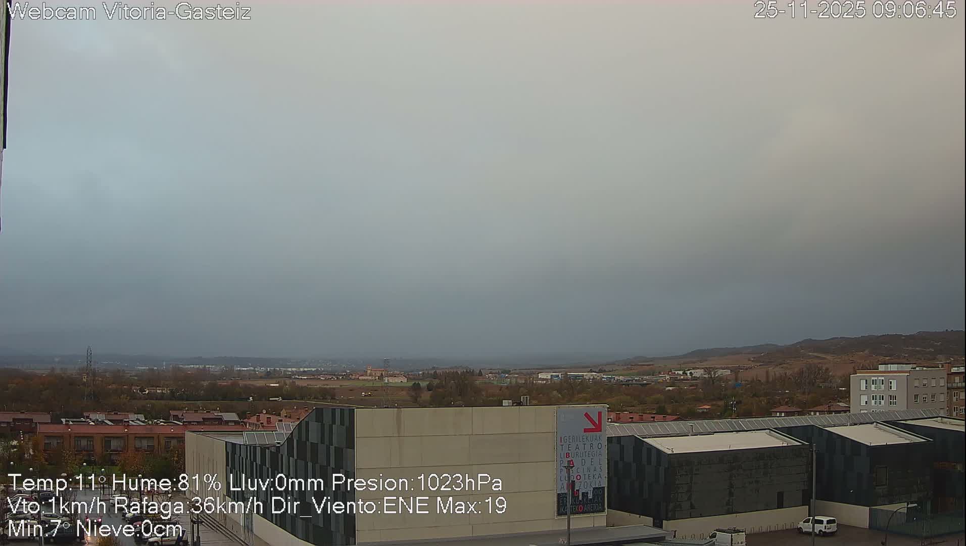 A wide-angle view reveals a modern urban landscape with distinctive dark-paneled buildings in the foreground, stretching towards residential areas and hazy hills under a uniformly grey, overcast sky.