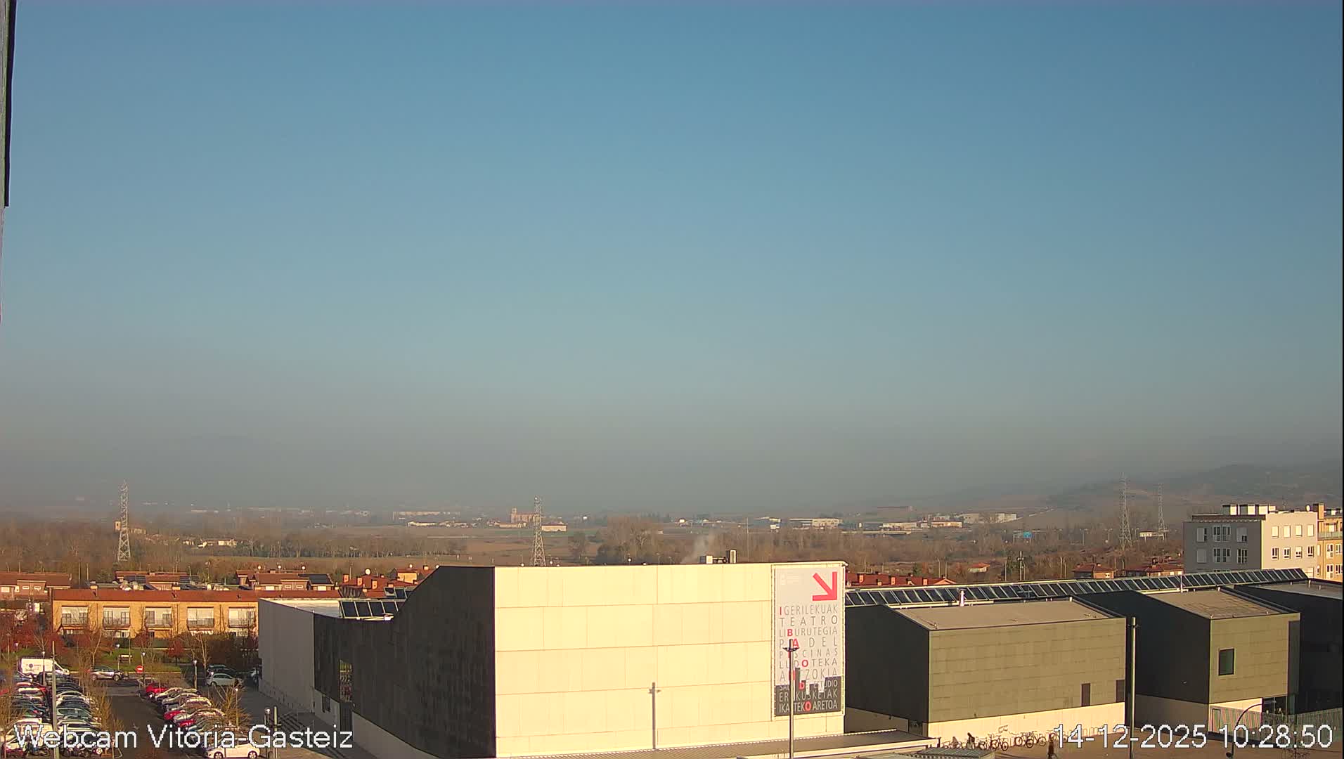 A wide view captures an urban landscape nestled in a valley, flanked by hills with some snow-dusted peaks, under a mostly cloudy sky with patches of blue.