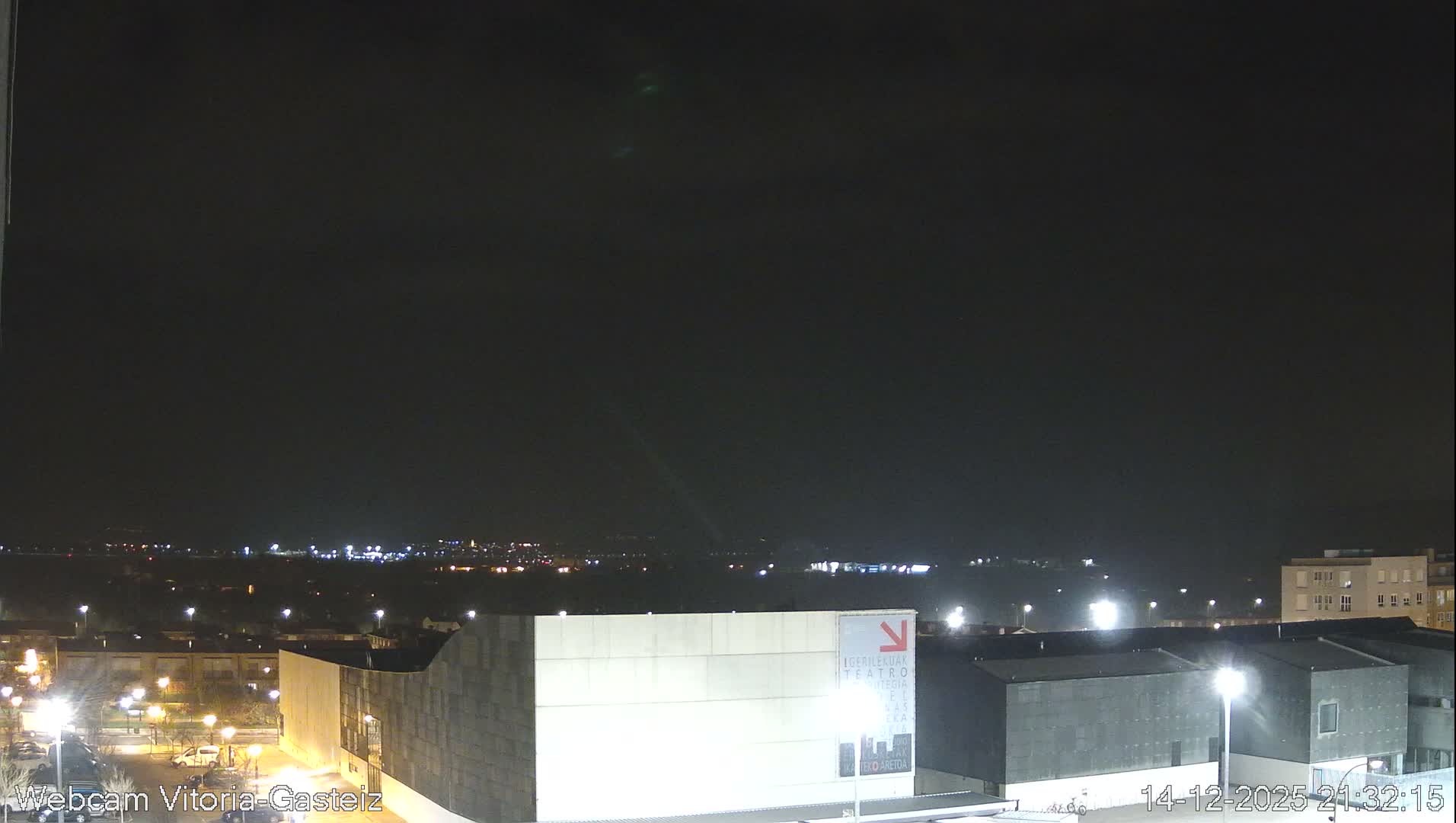 A wide view captures an urban landscape nestled in a valley, flanked by hills with some snow-dusted peaks, under a mostly cloudy sky with patches of blue.