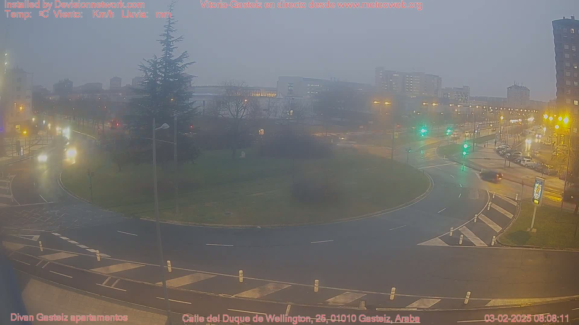 A wet roundabout in a city, shrouded in fog at night, with sparse traffic and buildings visible in the distance.