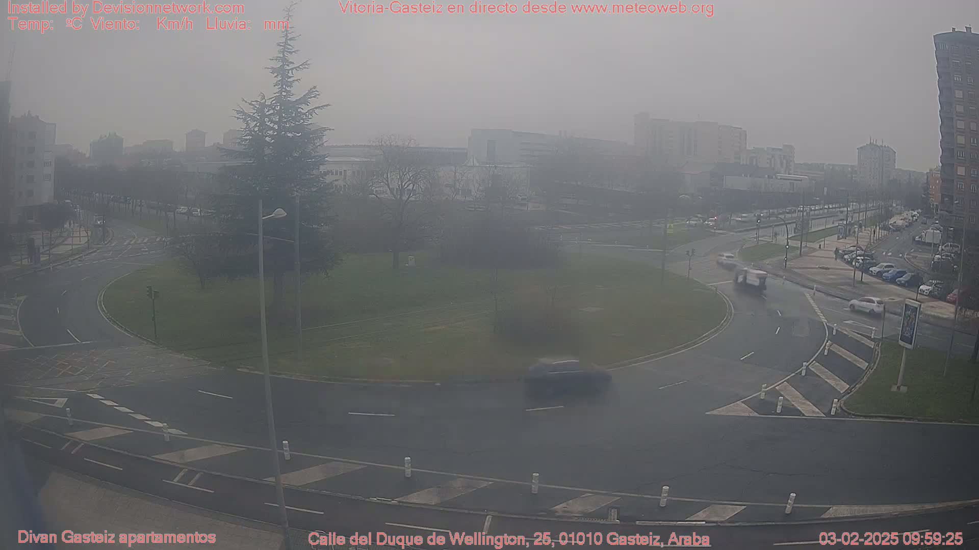 A hazy, overcast day shows a traffic circle with several cars, surrounded by buildings and trees.
