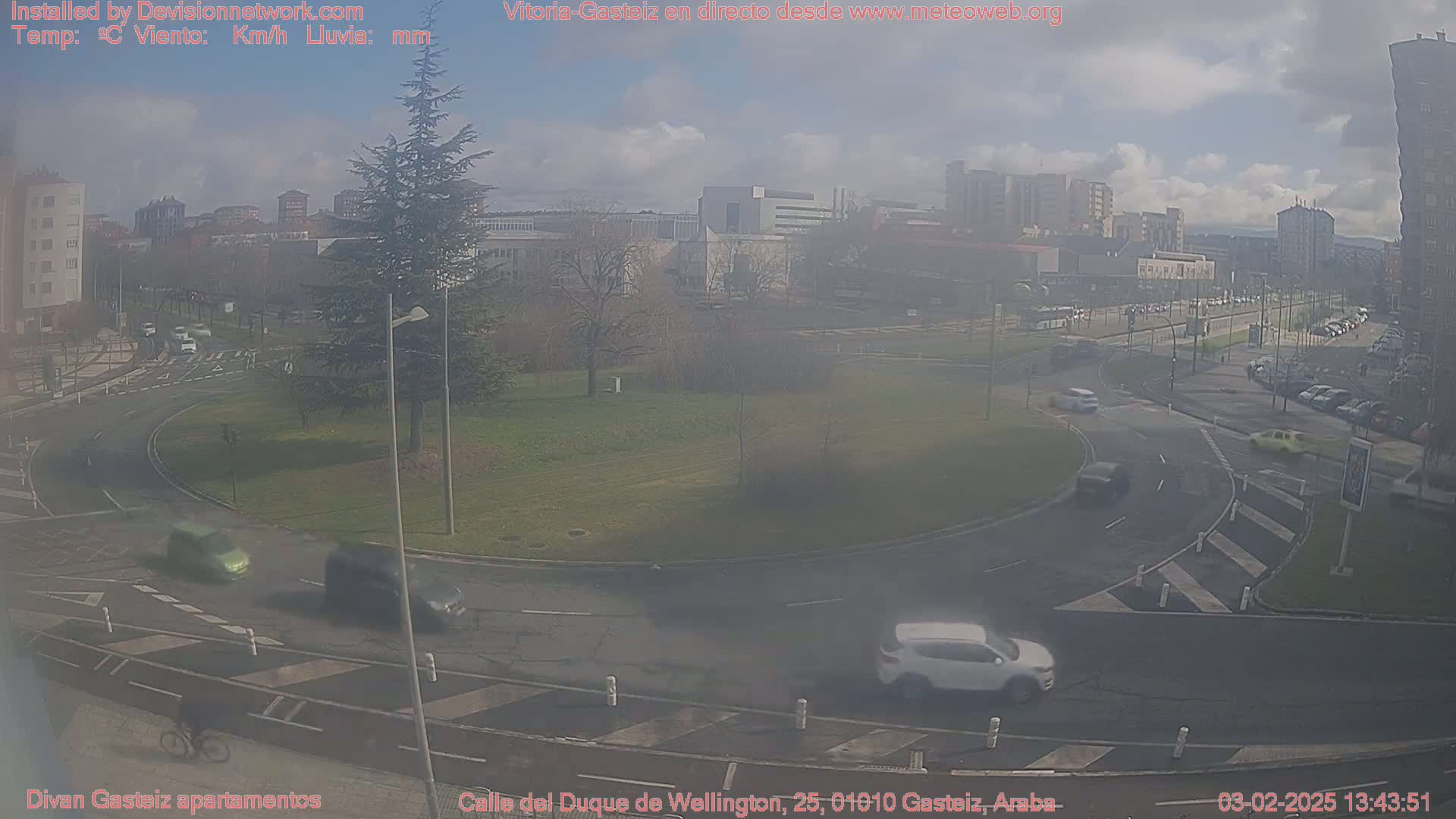 A hazy view of a roundabout with several cars and a cyclist, surrounded by buildings and a park under a partly cloudy sky.