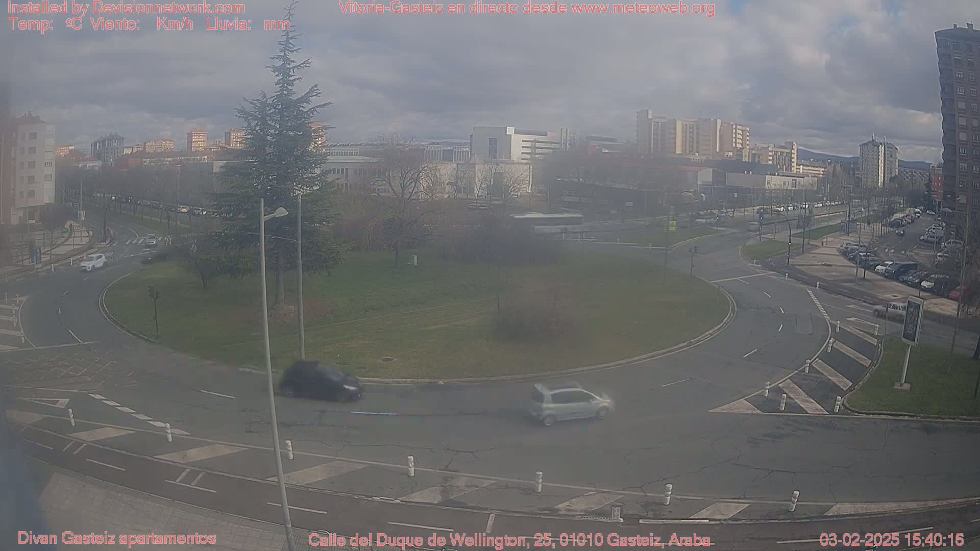 A hazy view of a roundabout in an urban area with several cars, surrounded by buildings and trees under a cloudy sky.