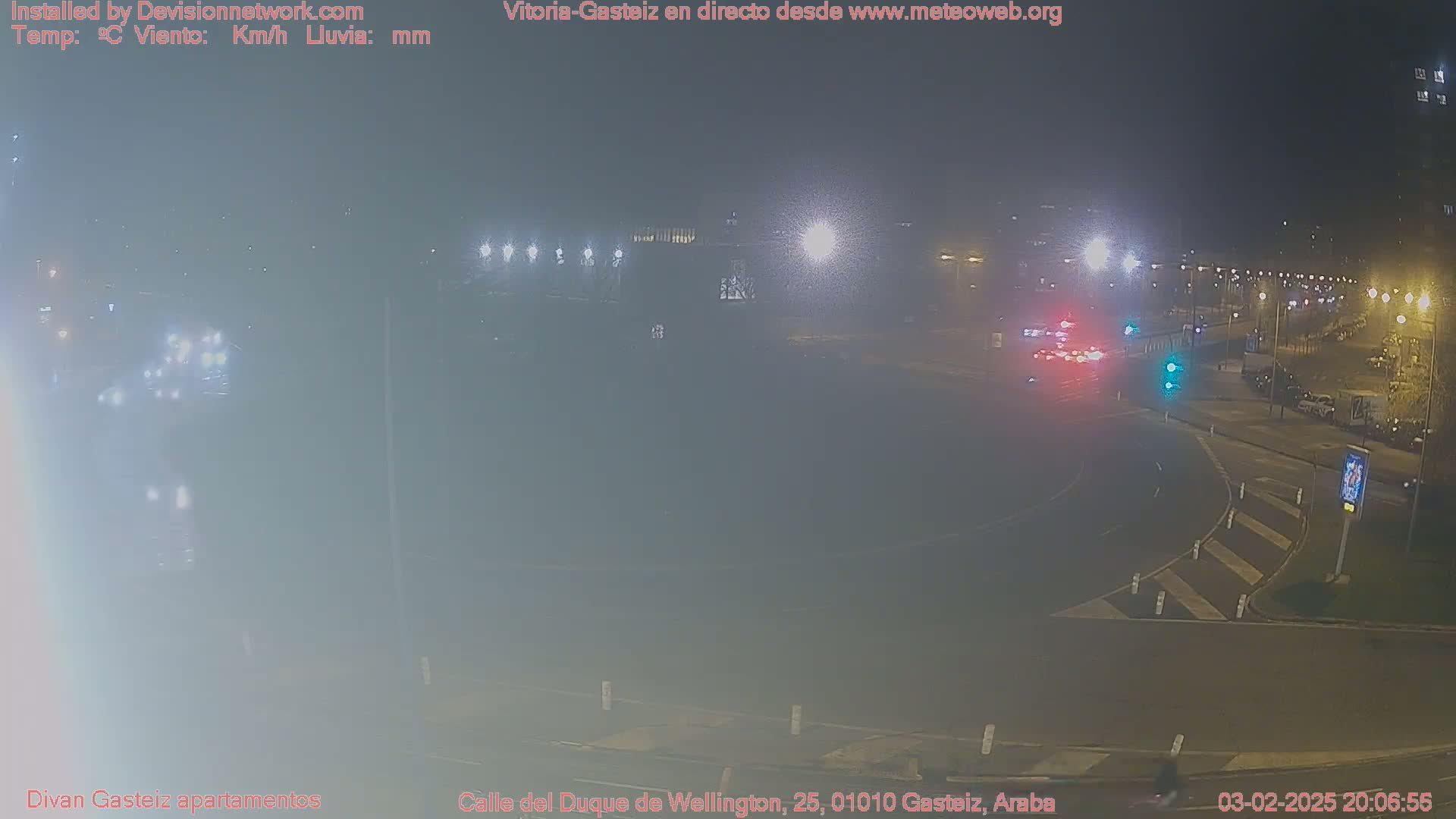 A nighttime view of a roundabout with several cars, streetlights, and buildings in the background, under hazy or foggy conditions.