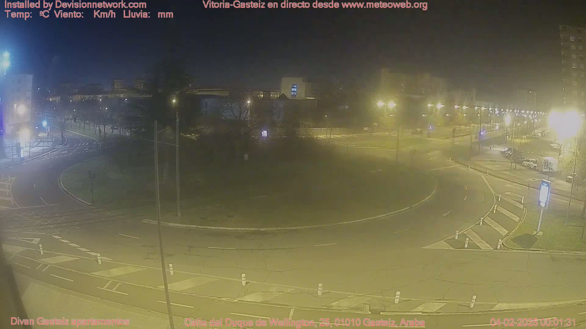 A nighttime view of a roundabout and surrounding streets, dimly lit with streetlights, showing some parked cars and a hazy atmosphere.
