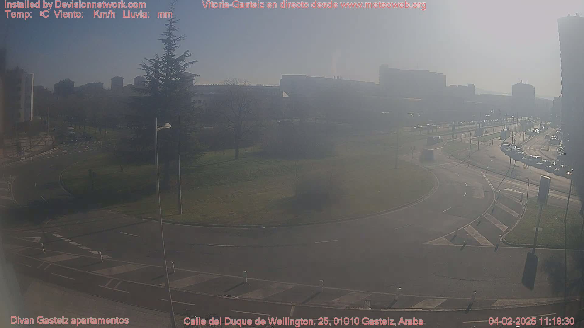 A hazy, sunny day reveals a roundabout and roadway intersection near a park and several buildings.