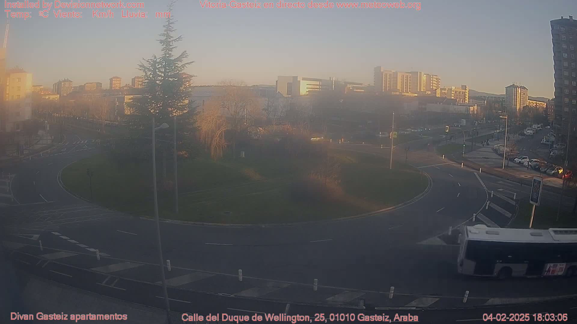 A hazy sunset illuminates a roundabout and city streets, with a bus driving past parked cars and buildings in the background.