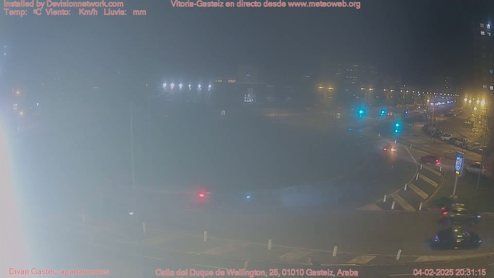 A foggy nighttime street scene shows a roundabout with several vehicles and traffic lights.