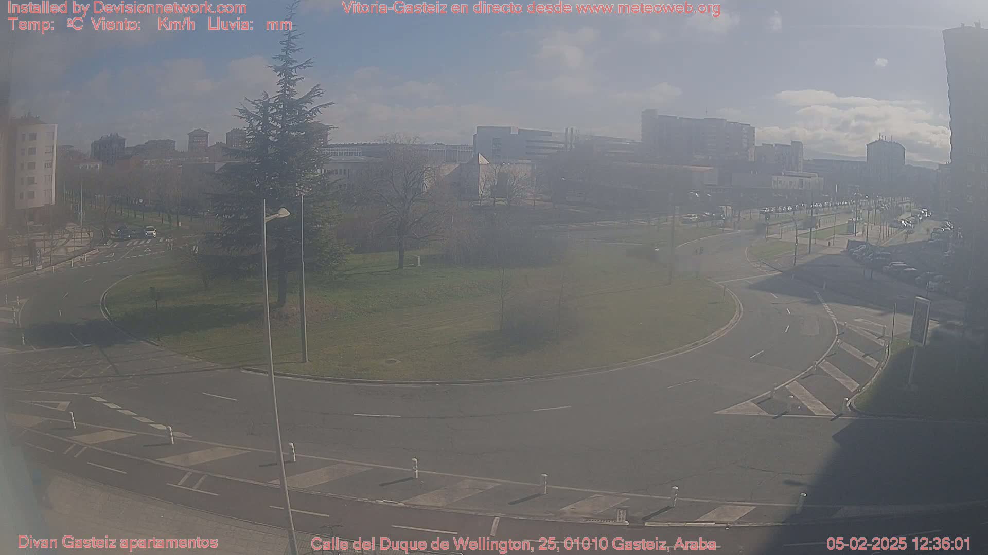 A hazy, sunny day reveals a city roundabout with surrounding buildings and a grassy area, featuring sparse traffic.