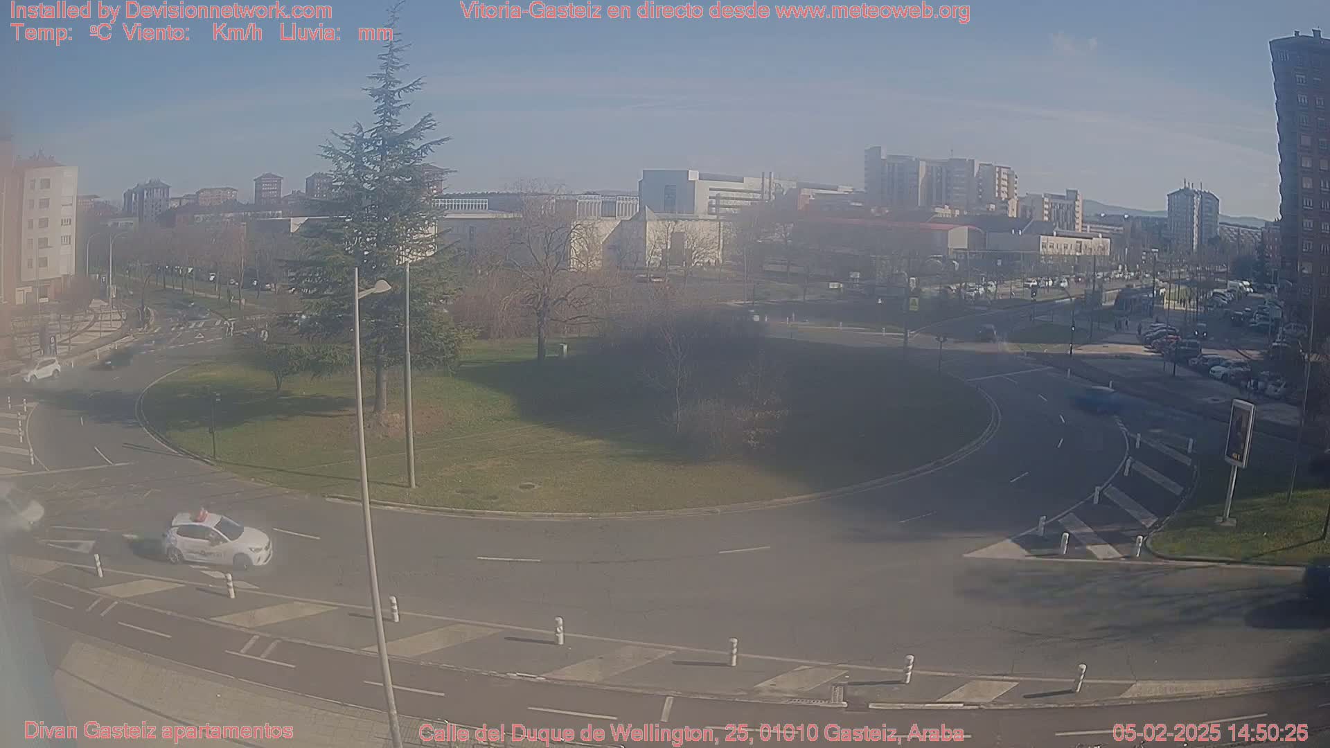 A sunny day reveals a roundabout in a city with several buildings in the background and a white car in the foreground.