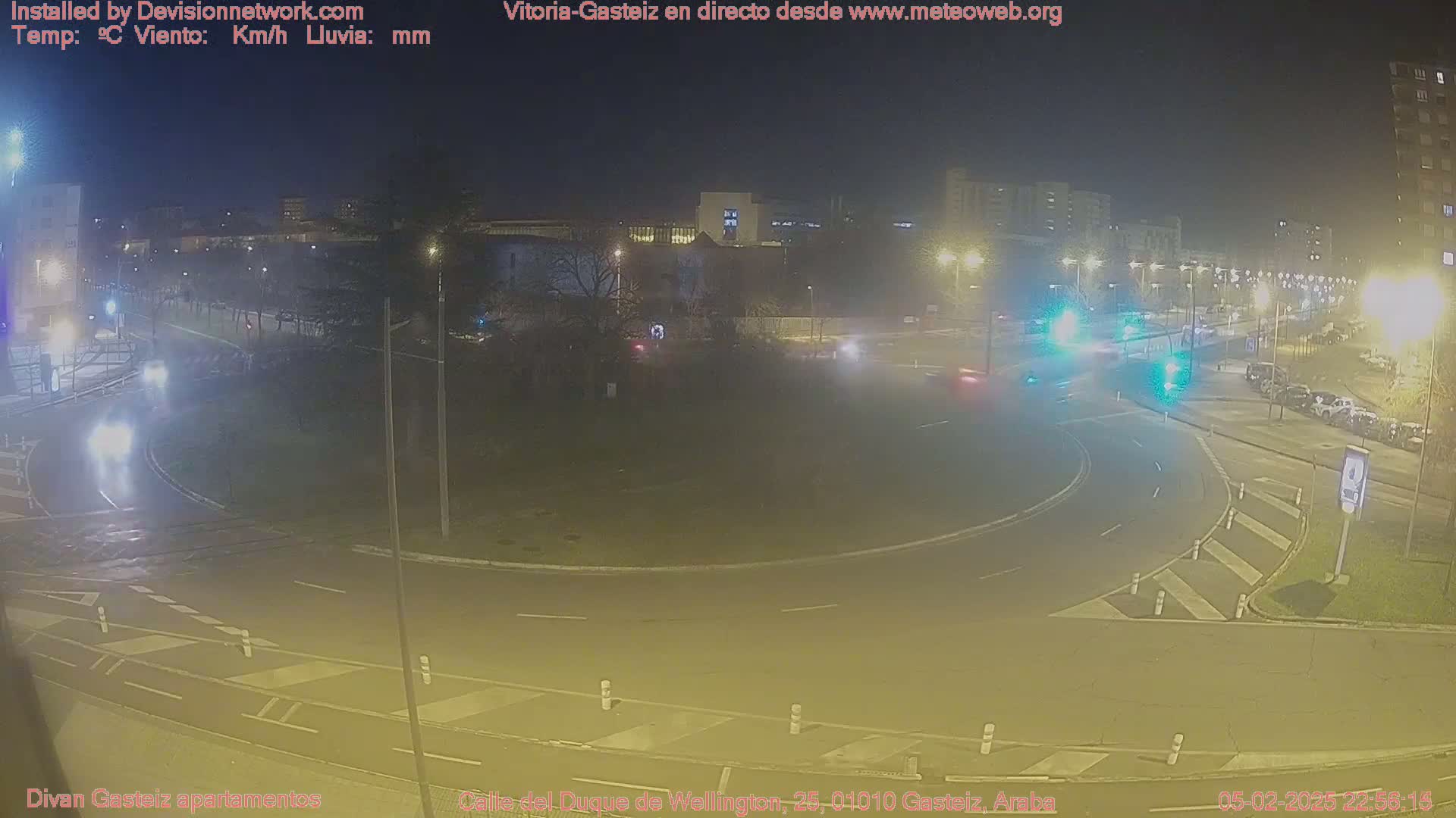 A nighttime, wet view of a roundabout with sparse traffic and streetlights illuminating the surrounding buildings and parked cars.