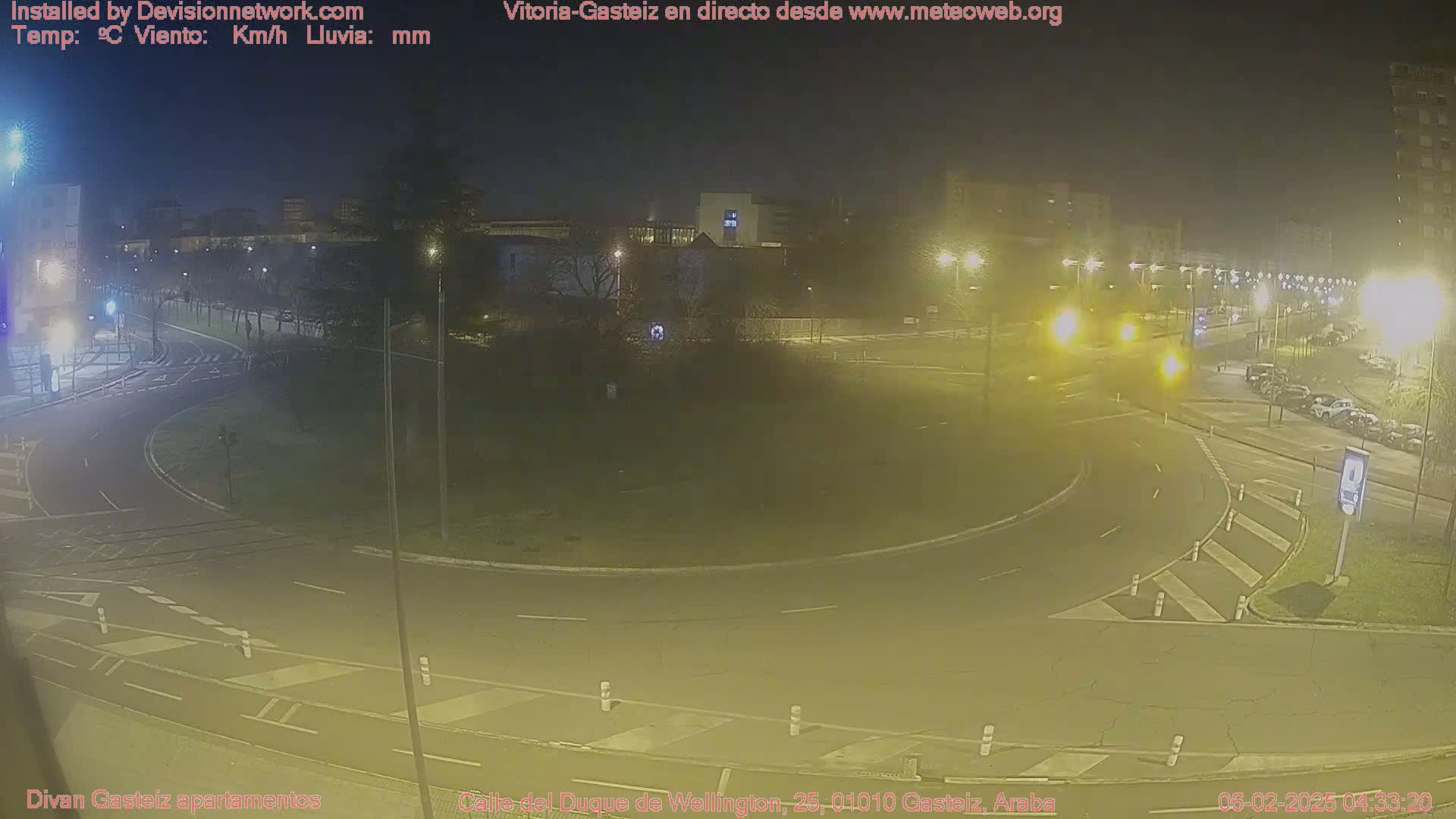 A nighttime view of a roundabout with sparse traffic, under partly cloudy skies illuminated by streetlights.