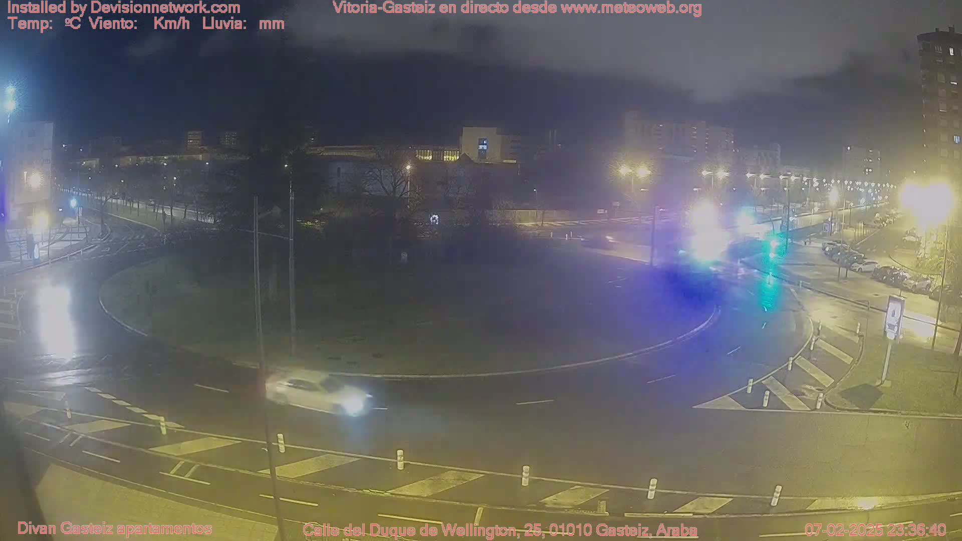 A nighttime view of a wet roundabout with cars driving through it, under streetlights, with buildings and trees in the background.