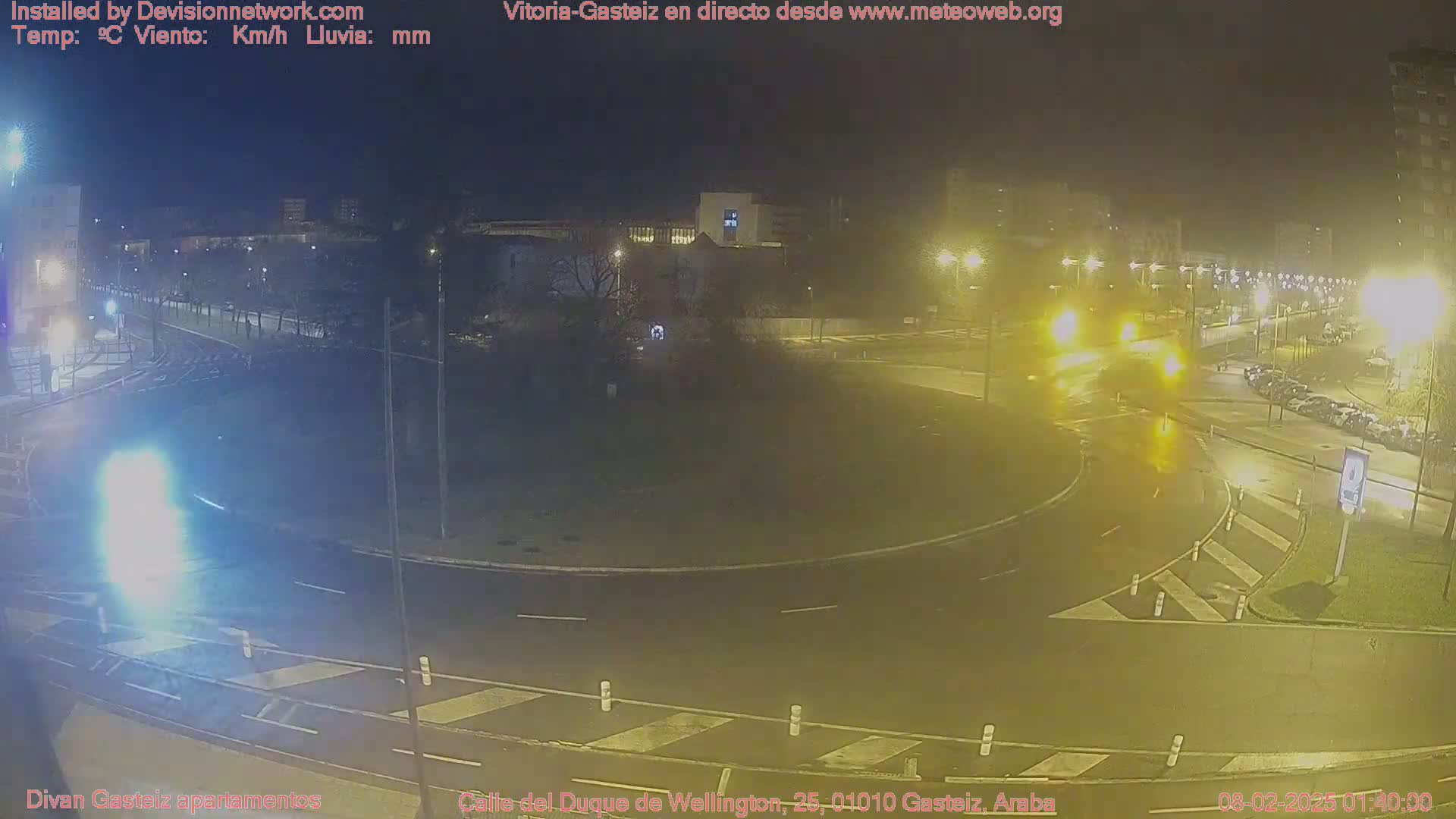 A nighttime view of a wet roundabout with cars and streetlights, under cloudy conditions.