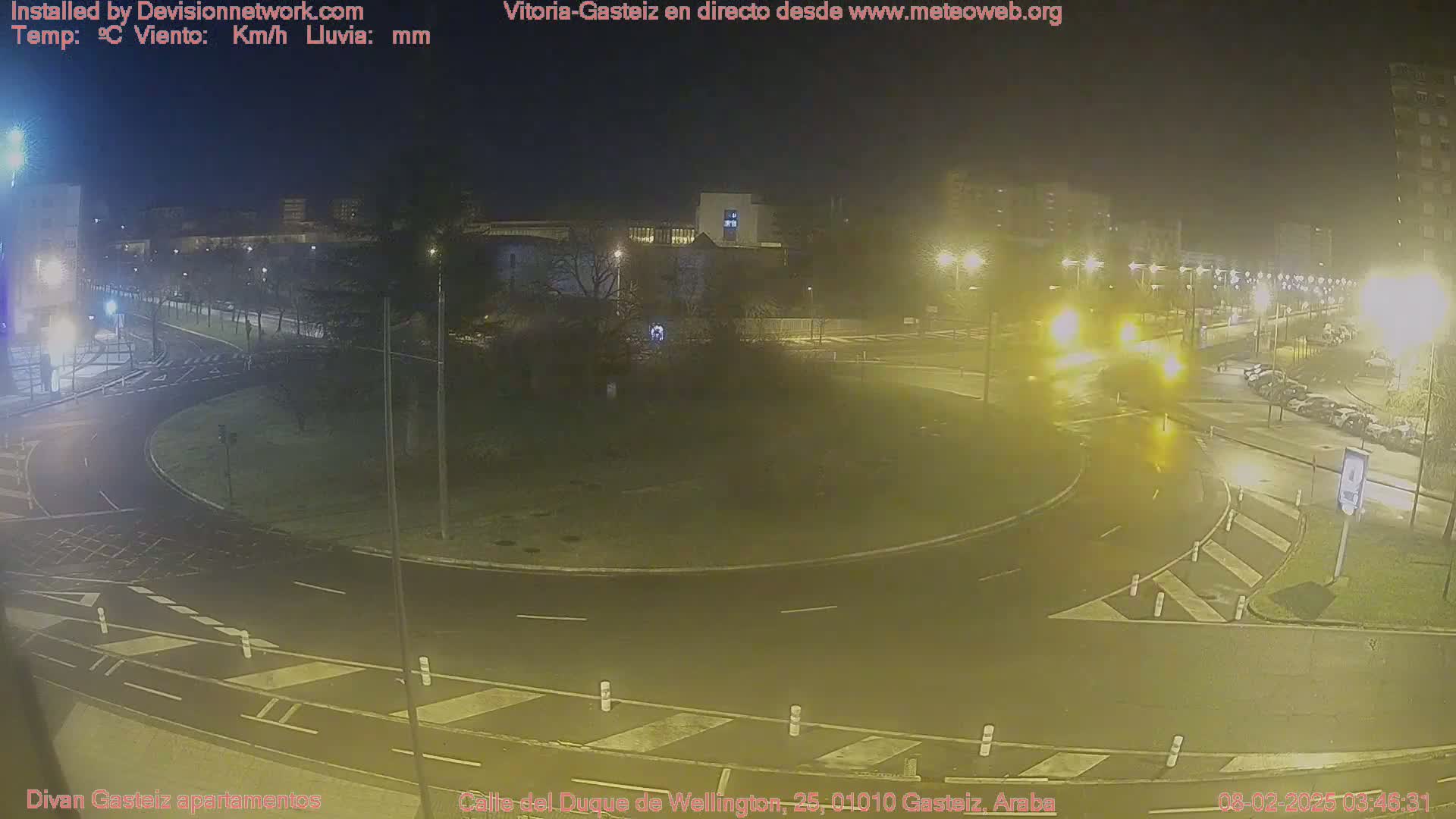 A nighttime view of a roundabout with cars, streetlights, and parked vehicles, under hazy conditions.
