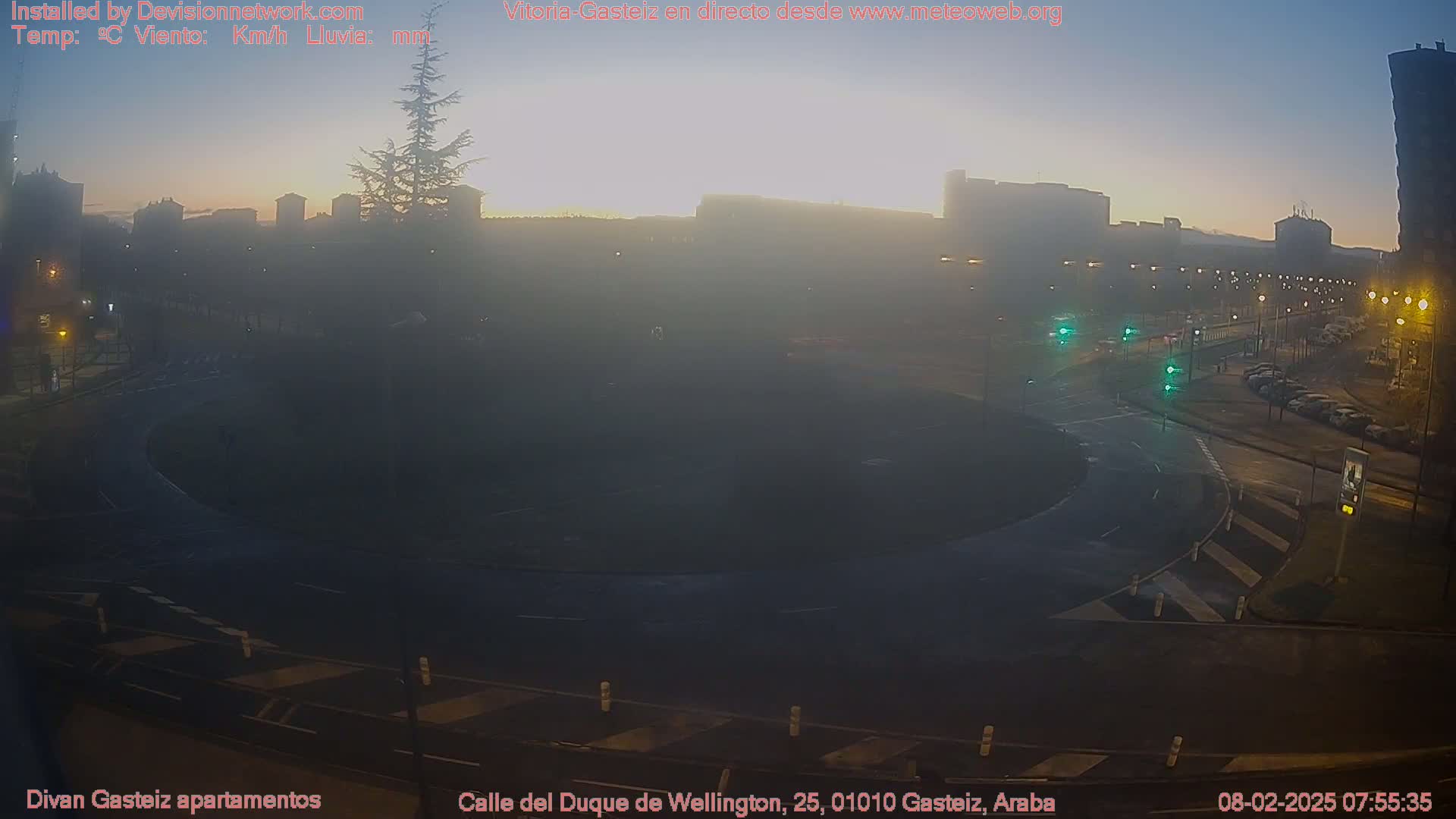 A hazy sunrise illuminates a city roundabout and surrounding buildings, with cars parked along the streets.
