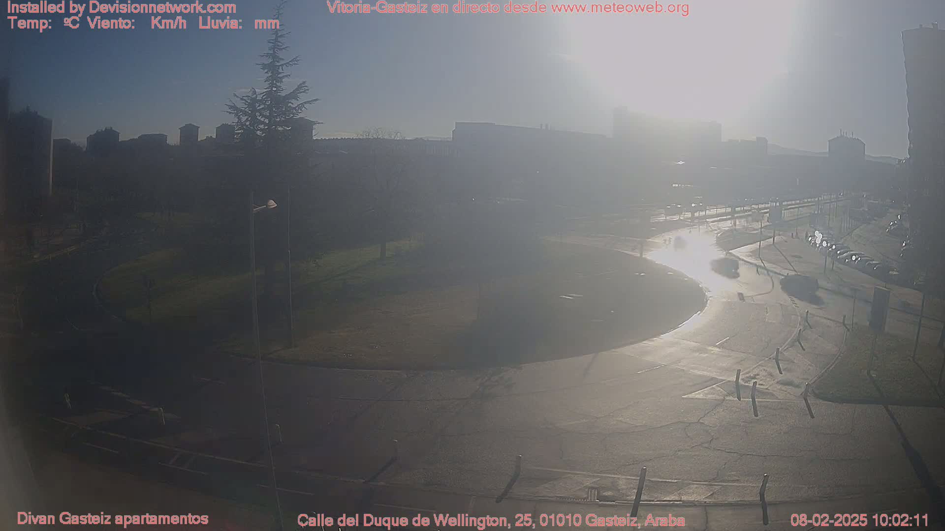 A hazy, sunny day reveals a city street scene with a roundabout, several parked cars, and buildings in the distance, featuring a large tree to the left.