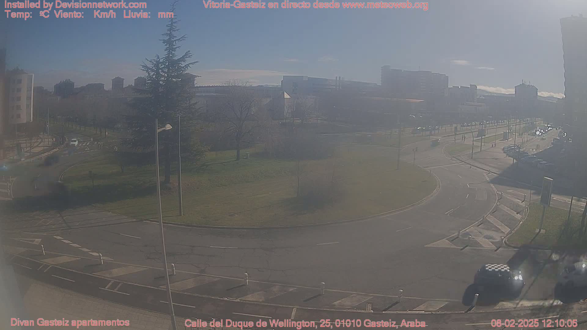 A hazy, sunny day reveals a roundabout and roadway with sparse traffic, surrounded by buildings and a green space.