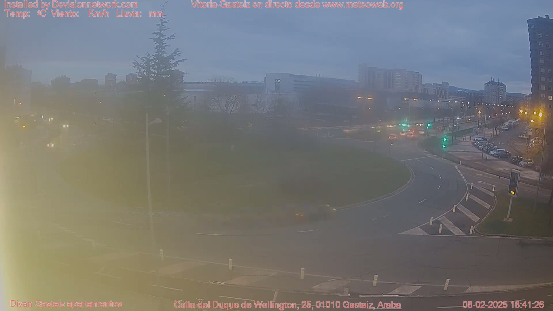A hazy dusk scene shows a roundabout with cars moving on a city street under a mostly cloudy sky.