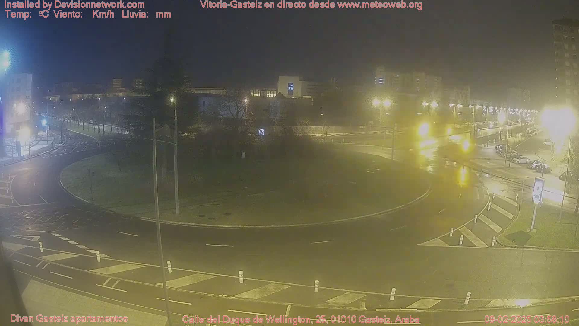 A wet roundabout at night, with cars driving through it and streetlights illuminating the surrounding area.