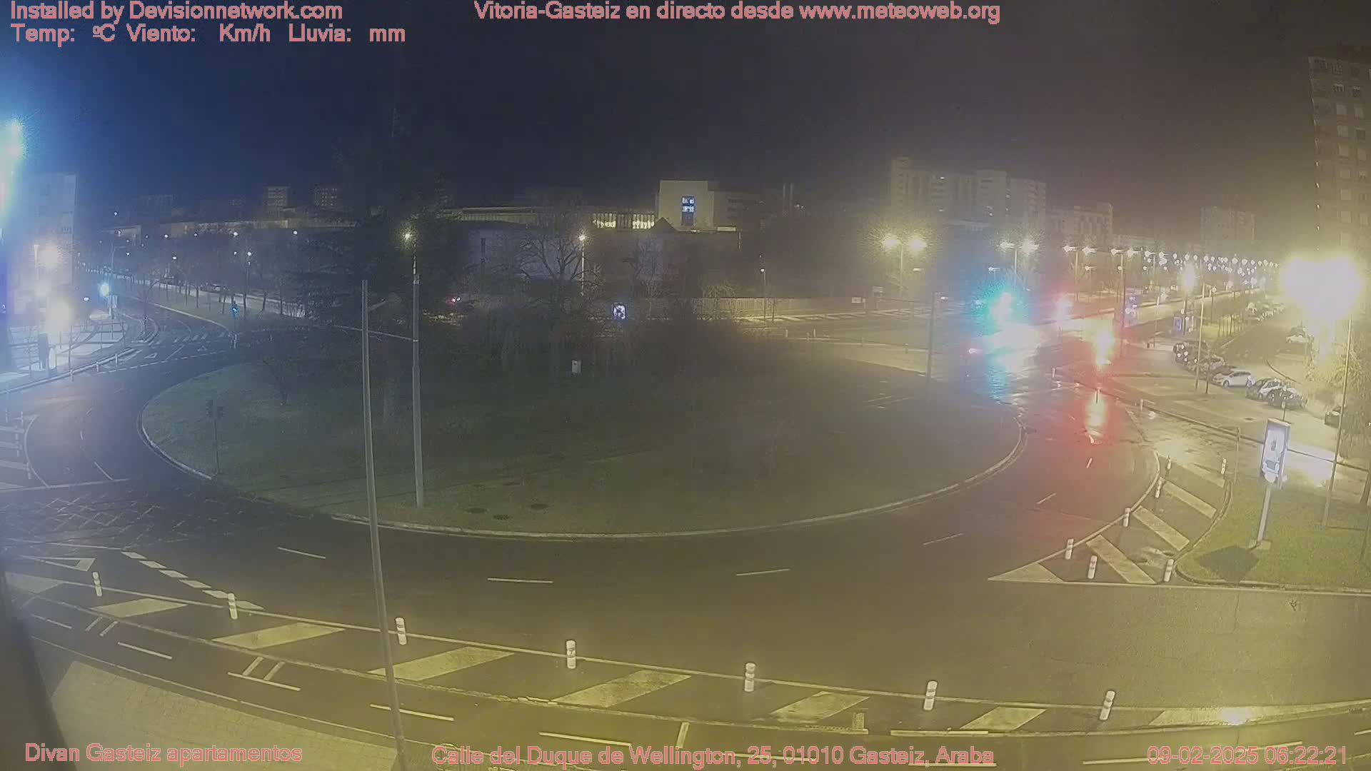 A wet roundabout at night with several cars and streetlights illuminated.