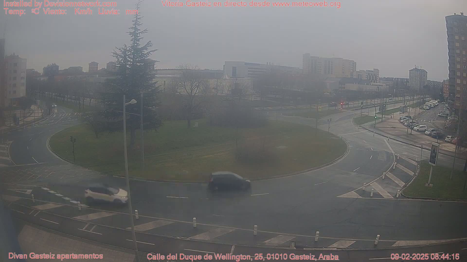 A hazy, overcast day shows a traffic circle with several cars, surrounded by buildings and trees.