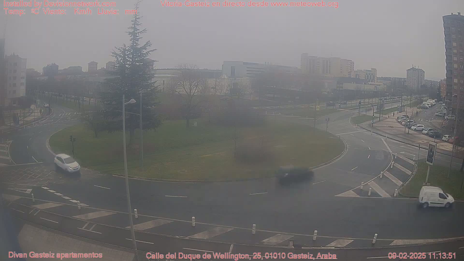 A hazy, overcast day reveals a roundabout intersection with several cars and a van in a city setting.