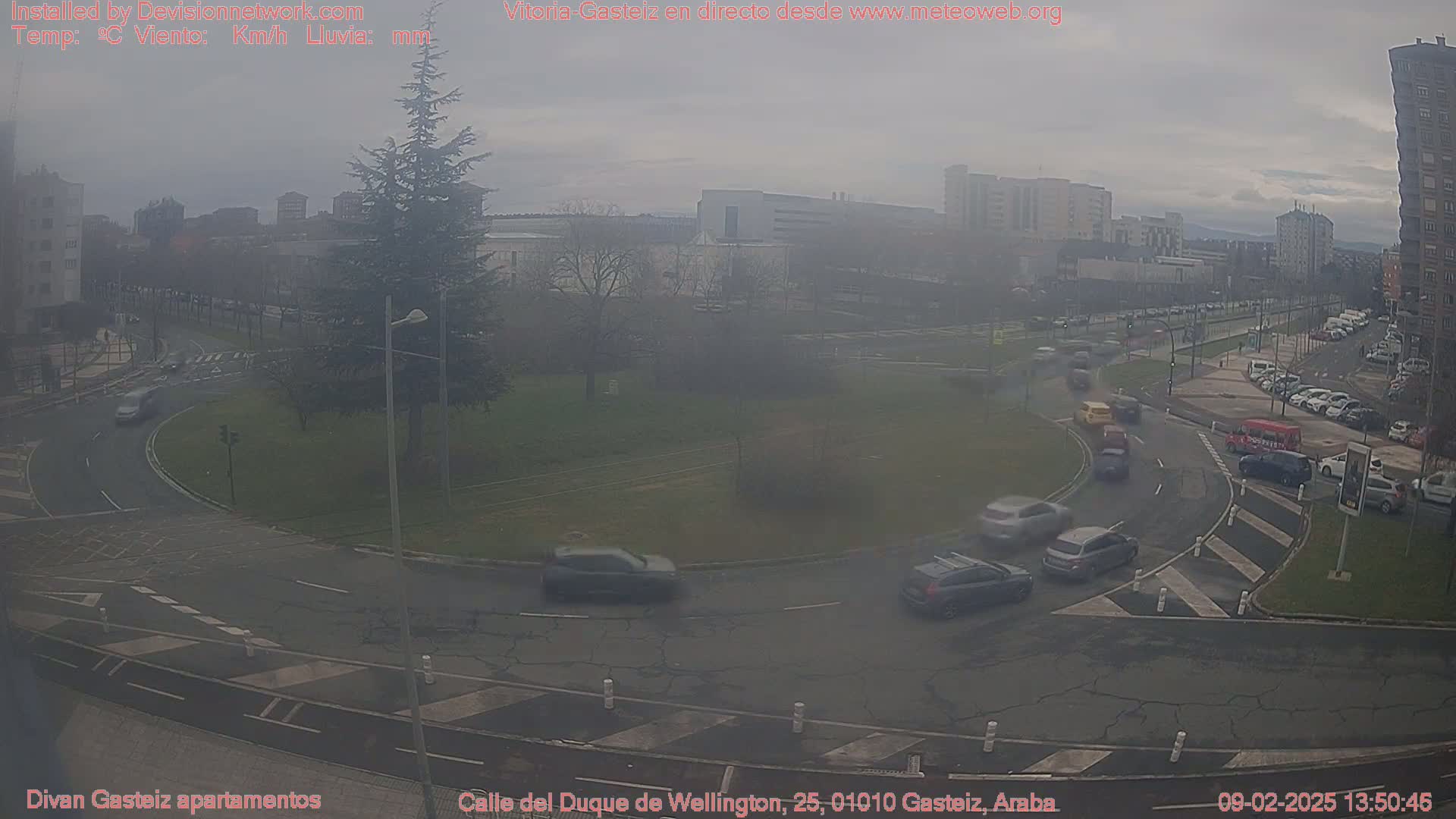 A hazy, overcast day shows a roundabout with several cars moving through it, surrounded by buildings and trees.