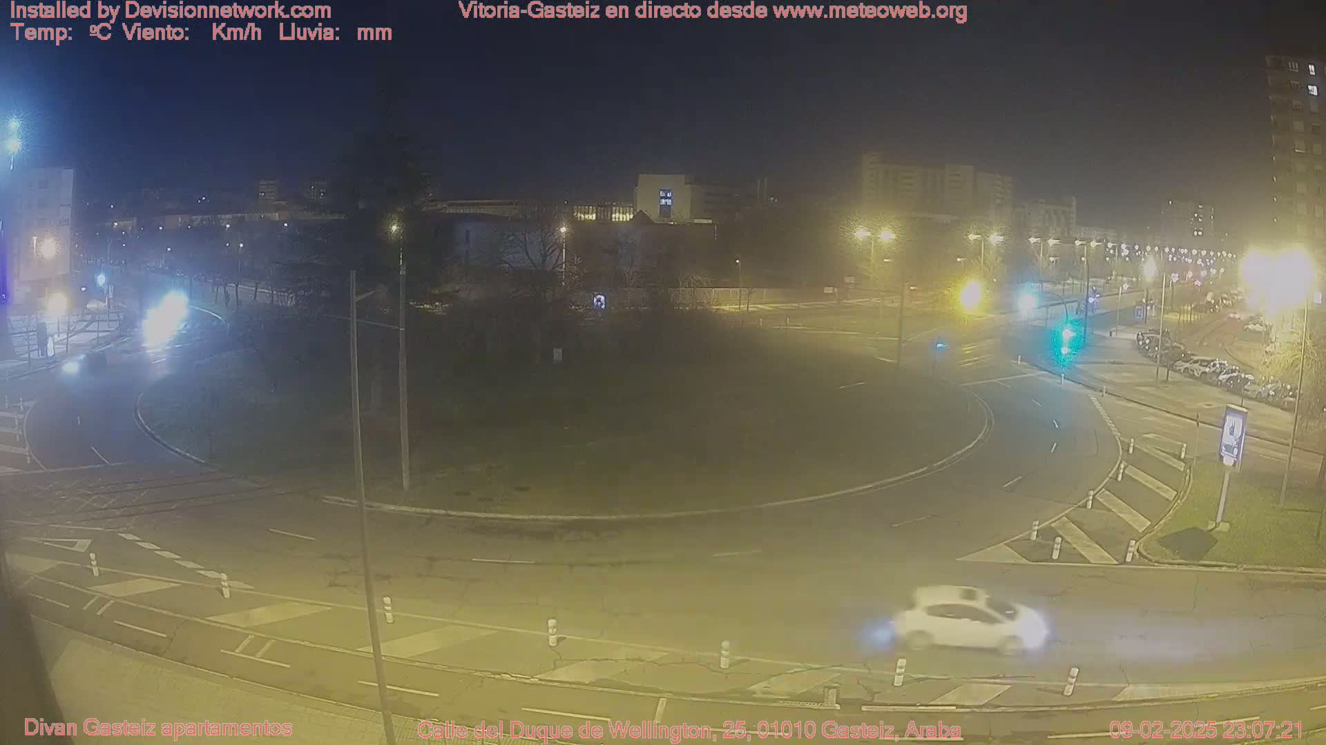 A nighttime view of a roundabout with a few cars, surrounded by buildings and streetlights under partly cloudy skies.