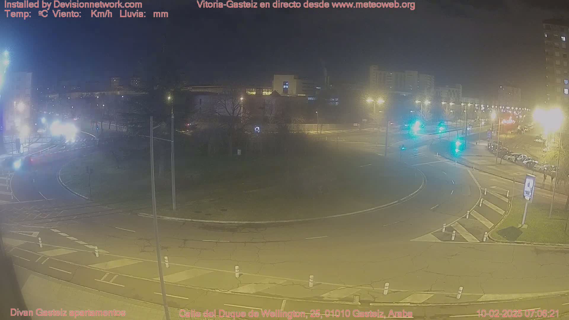 A nighttime view of a roundabout in a city with several cars driving through, streetlights illuminating the area, and buildings visible in the background.