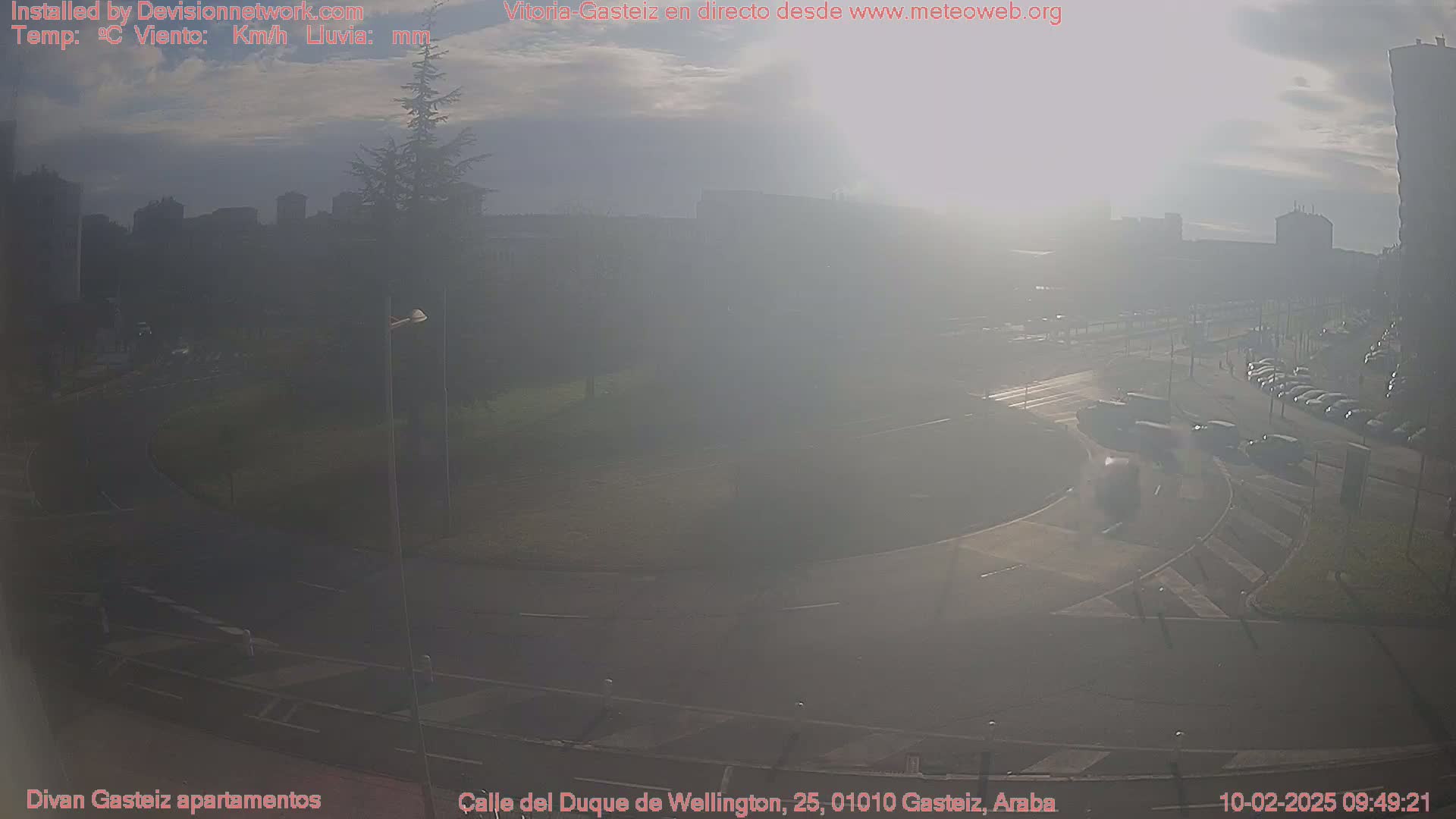A hazy, sunny view of a roundabout and city streets with several cars and buildings in the distance.