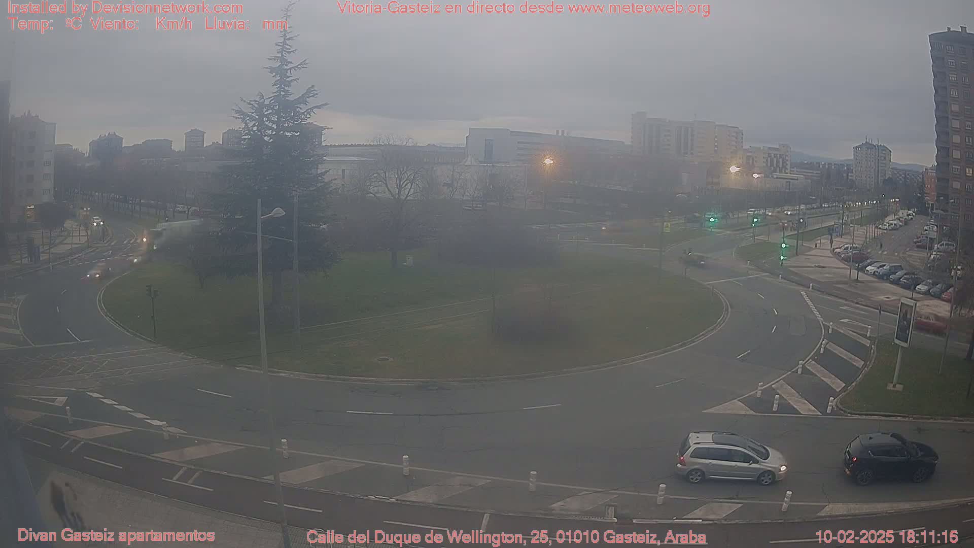 A hazy, overcast day reveals a roundabout with several cars, surrounded by buildings and trees.