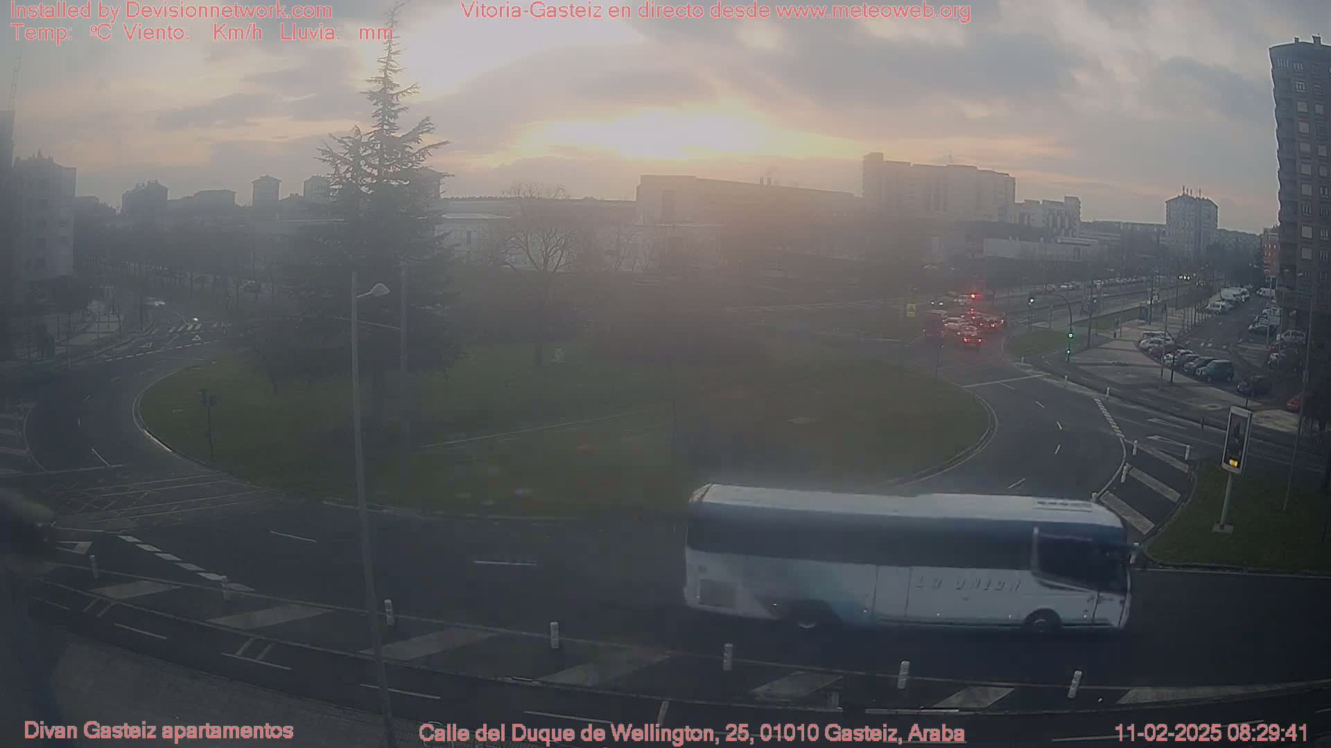 A hazy sunrise illuminates a roundabout and surrounding city streets with a bus traveling on the road.